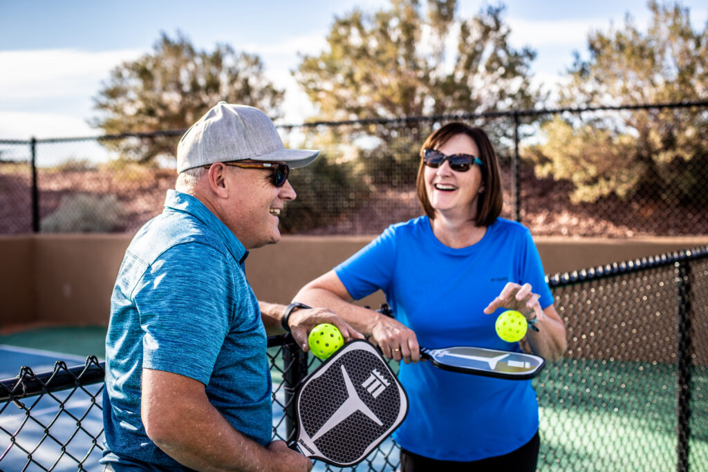 A smiling man and woman chatting on an outdoor pickleball court while holding paddles and neon balls, showcasing active amenities found at large family reunion vacation rentals.