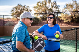 A smiling man and woman chatting on an outdoor pickleball court while holding paddles and neon balls, showcasing active amenities found at large family reunion vacation rentals.